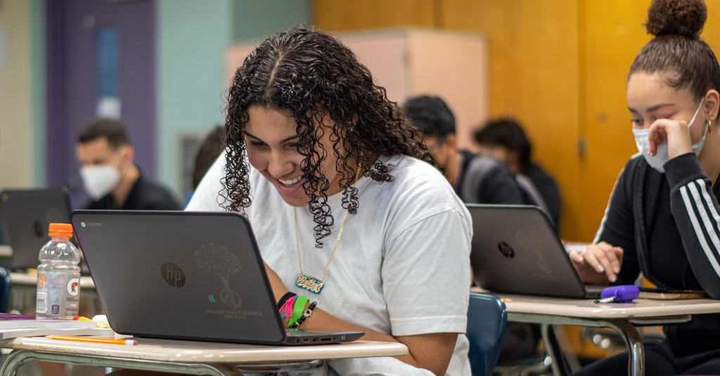 student smiling  and working on her laptop in a classroom with other students 