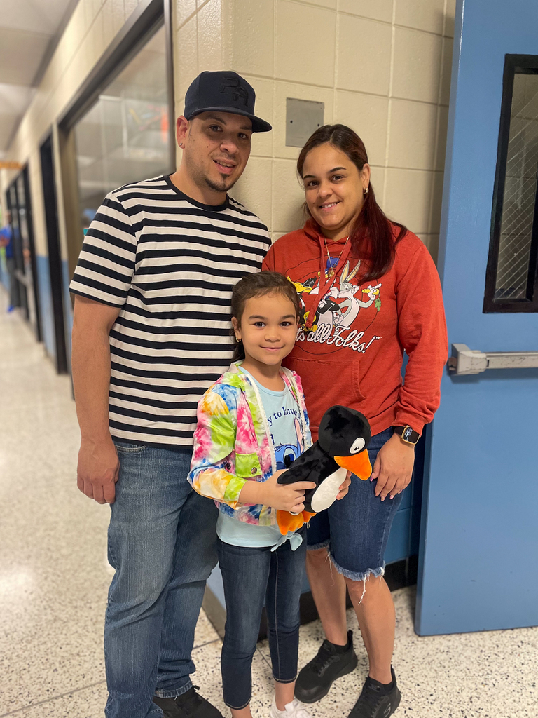 A family of three smiling as they pose with young student holding a stuffed animal.