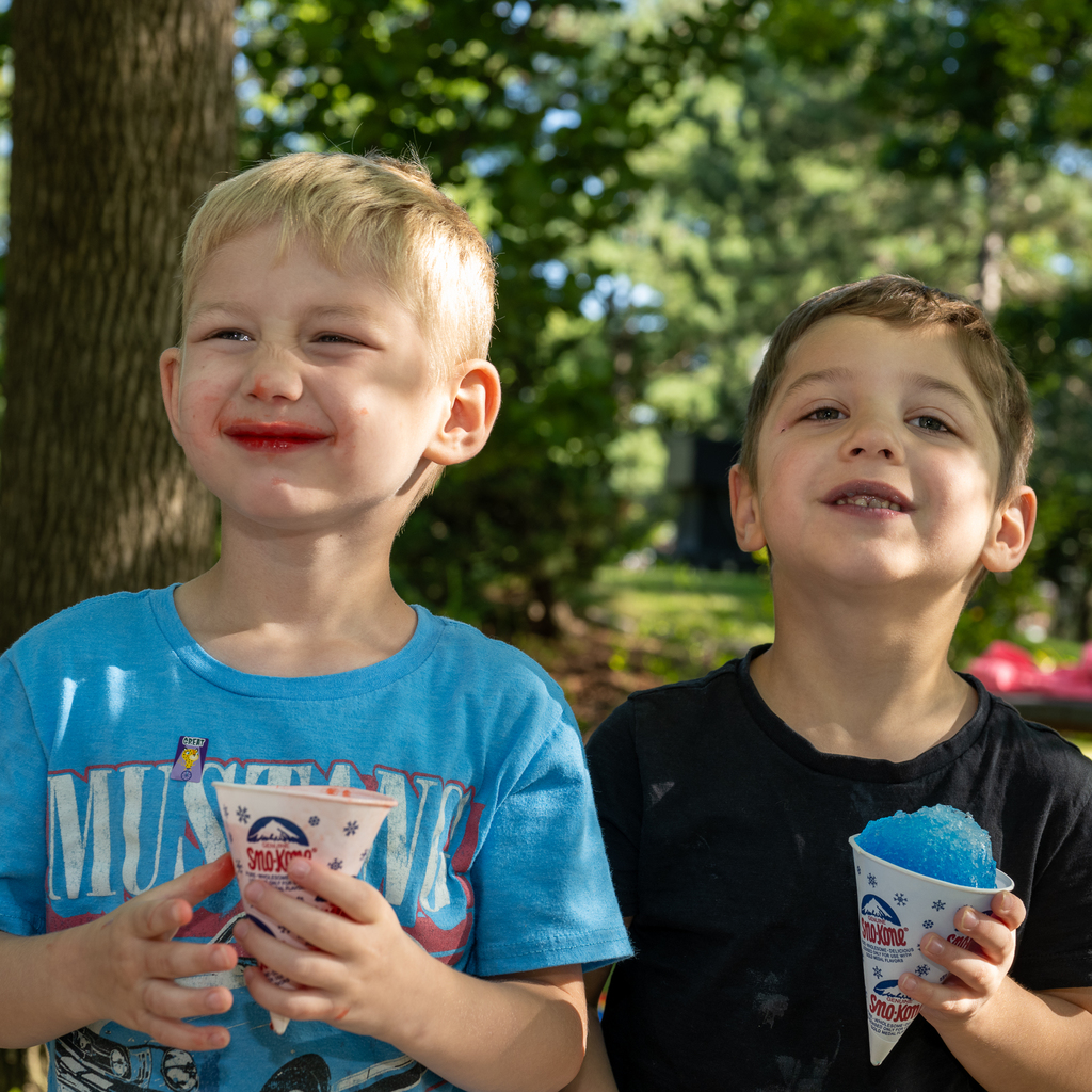 Two young boys next to each other smiling as they enjoy a snow kone. 